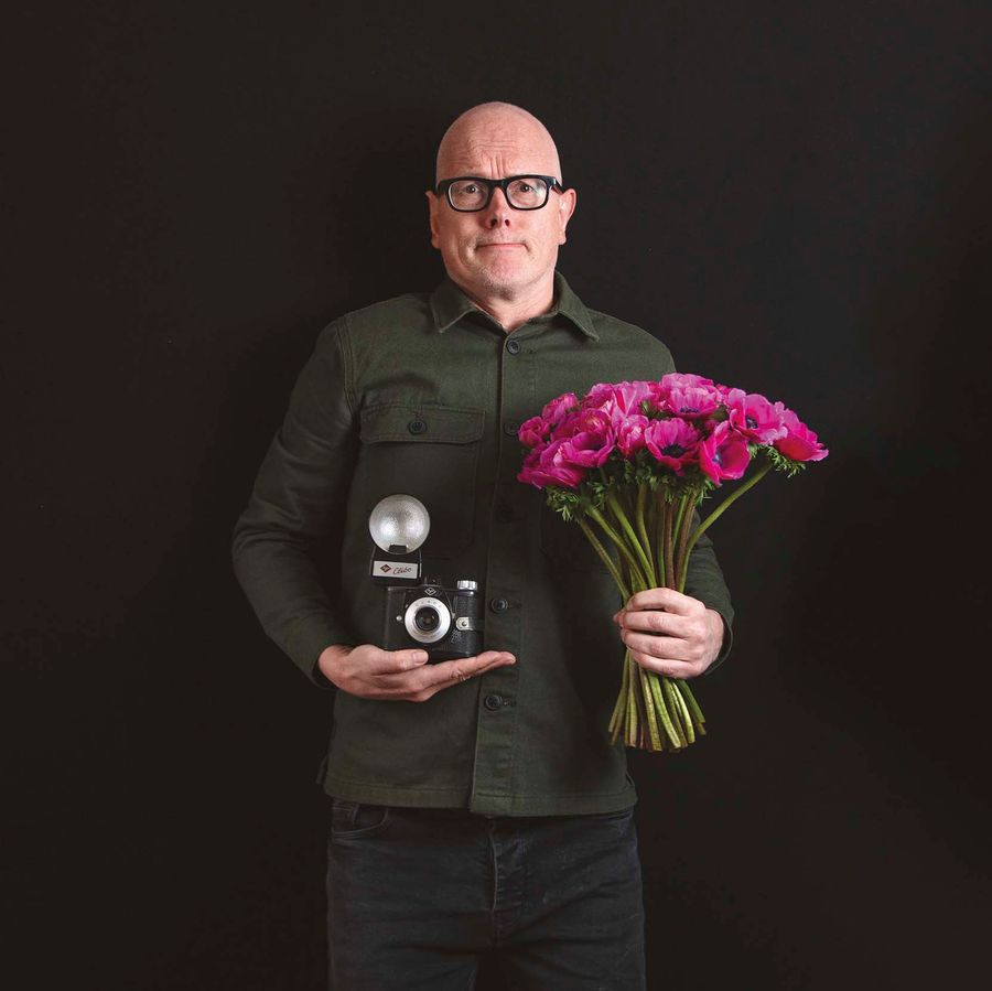Kees heeft een bos bloemen en een camera vast. Hij staat voor een zwarte achtergrond.