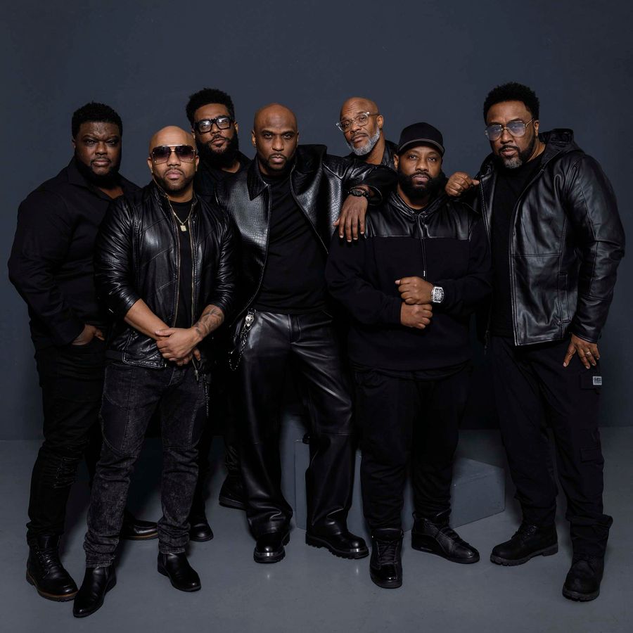 Groepsfoto van Naturally7 in zwarte kleding, poserend tegen een donkere achtergrond.