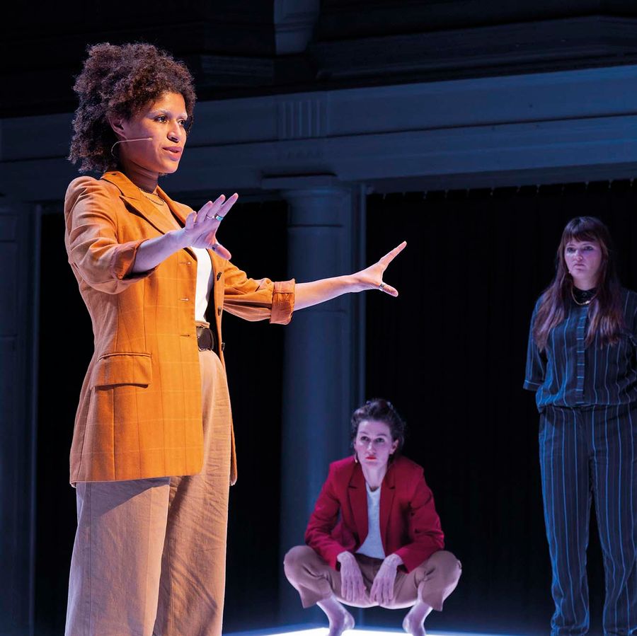 Een vrouw in een oranje pak is aan het praten. Drie vrouwen achter haar luisteren.