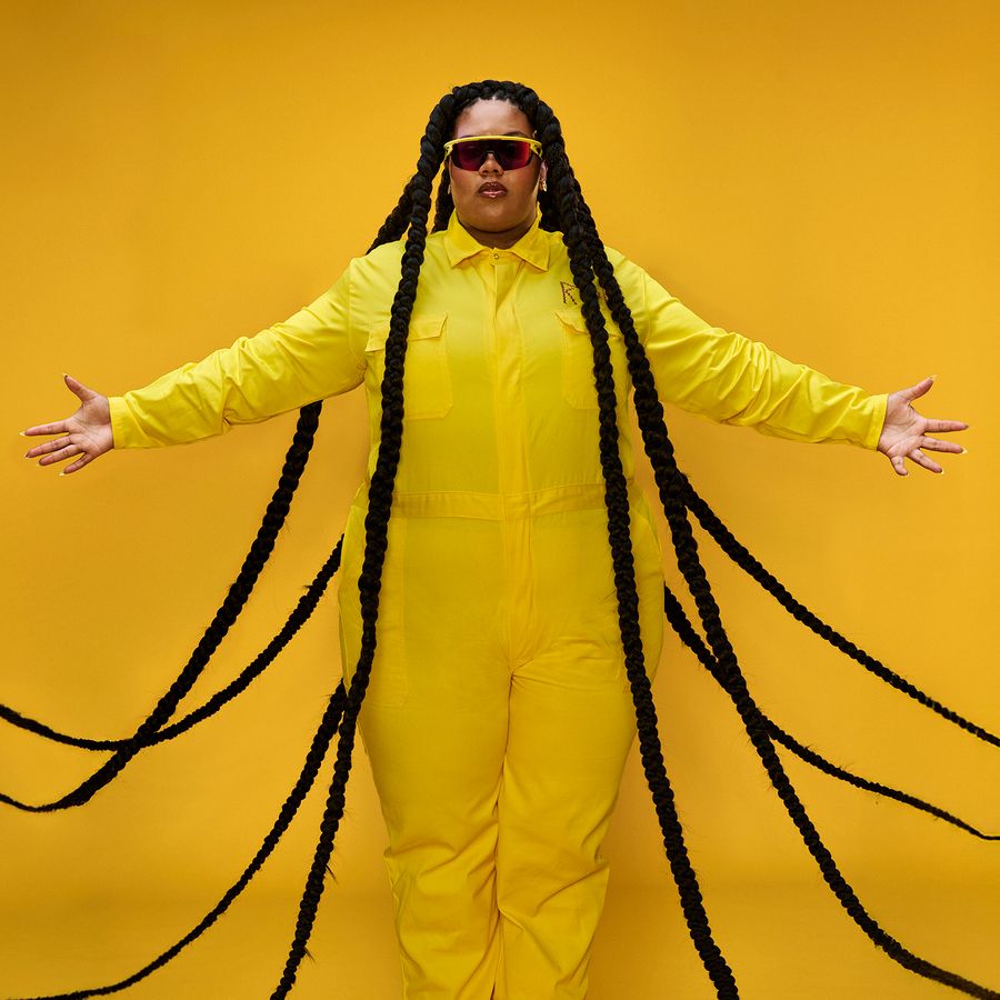 Een vrouw in een geel pak voor een gele achtergrond. Ze heeft haren die tot de grond komen.