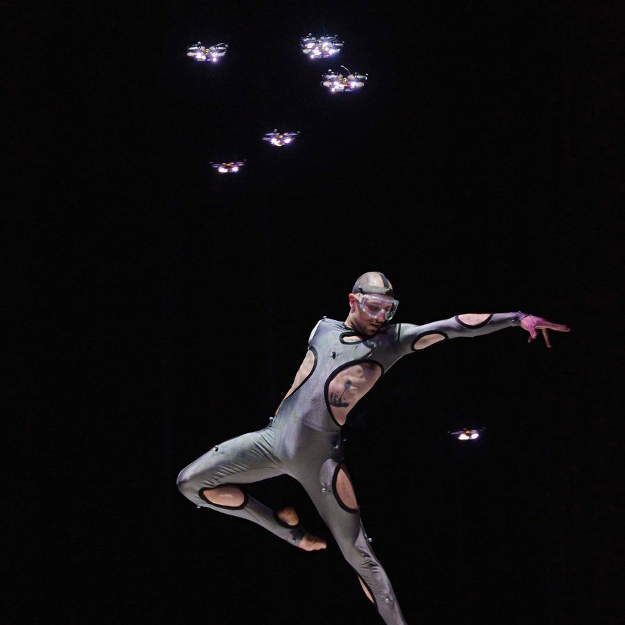 Een man met een bril op en een strak pak aan met gaten er in danst op het podium.