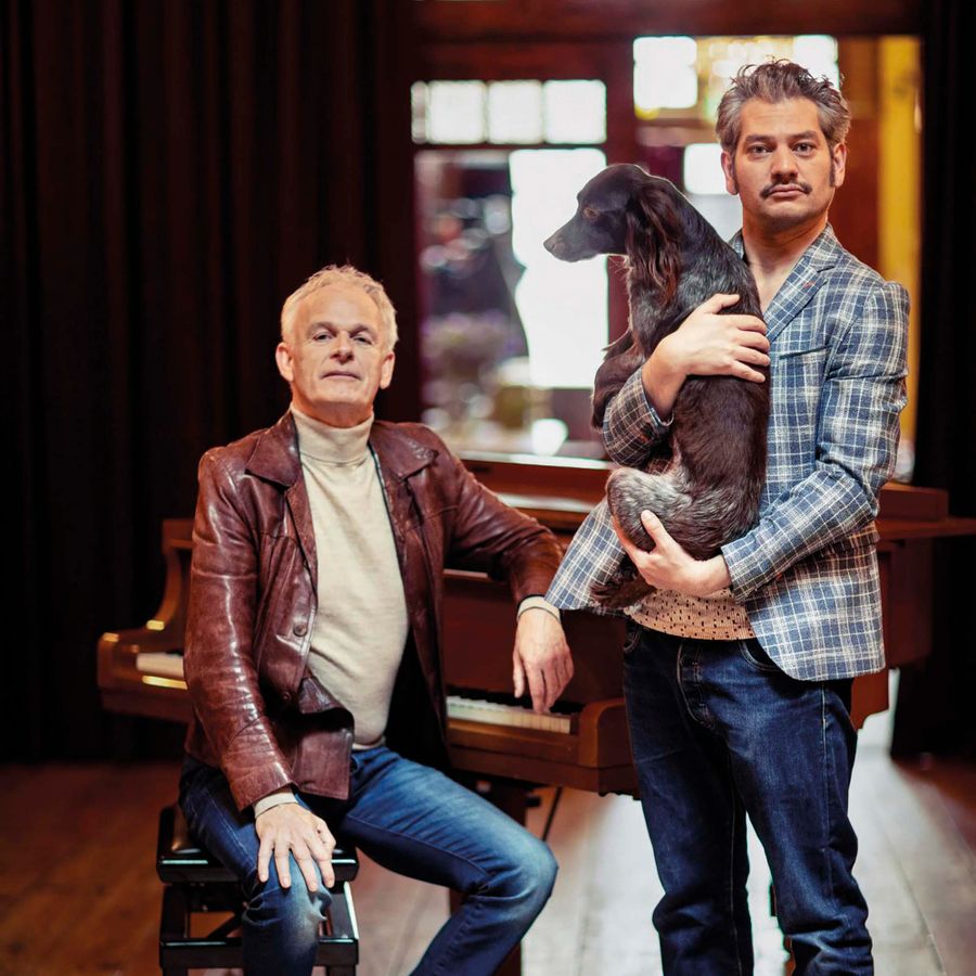 Een man zit op een stoel. Naast hem staat een man met een hond in zijn armen.