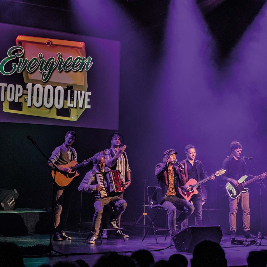 Een zanger zit op een krukje op het podium. De band staat om hem heen.