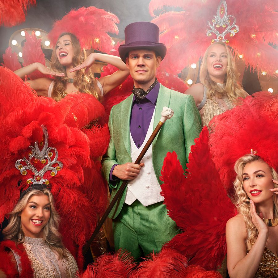 Tim in showkostuum tussen showgirls met rode veren in glamoureuze theatersetting.