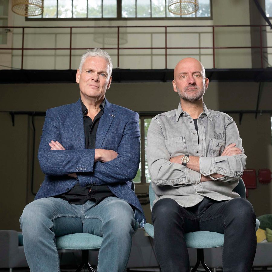 Joep en Rob zitten naast elkaar op een bankje met de armen over elkaar.