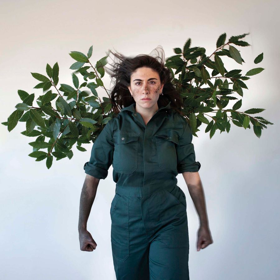 Een vrouw in een groen pak met modder op haar gezicht. Er hangt een tak met bladeren achter haar.