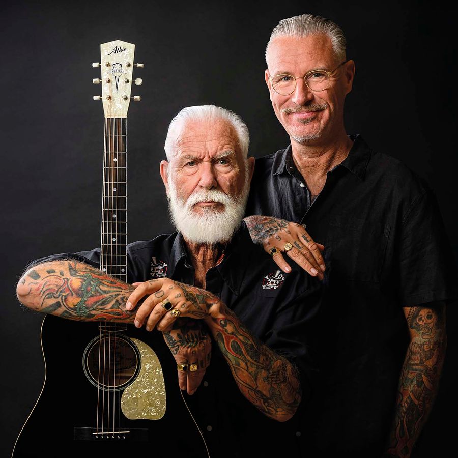 Twee mannen staan naast elkaar met een zwarte achtergrond. een leunt op een gitaar.