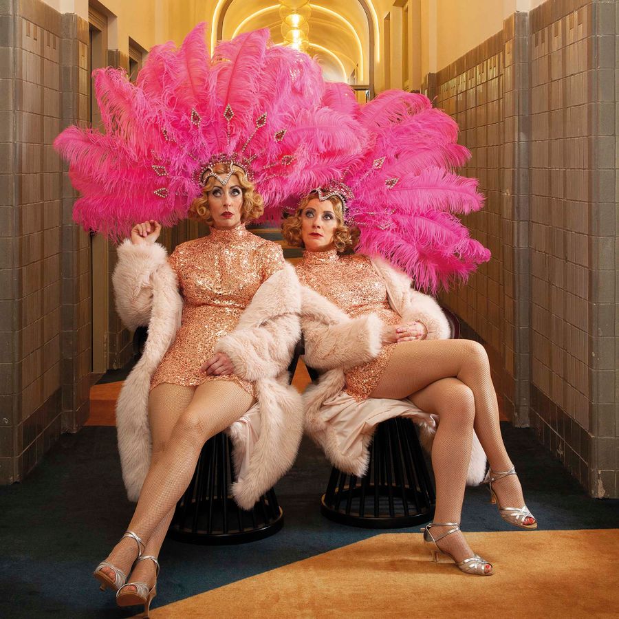 Plien en Bianca in glitterjurken en grote roze verentooi poseren in een stijlvol verlichte gang.