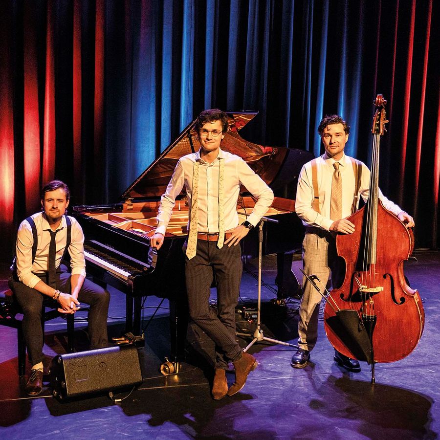 Drie mannen poseren voor een piano. Ze dragen en witte blouse met een stropdas.