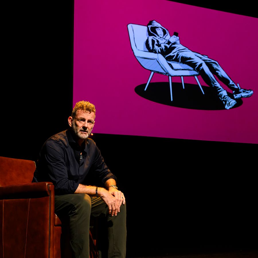 Kluun zit op een stoel op het podium met achter hem een projectie van een puber in stoel op felroze achtergrond, uit voorstelling Help, ik heb een puber!