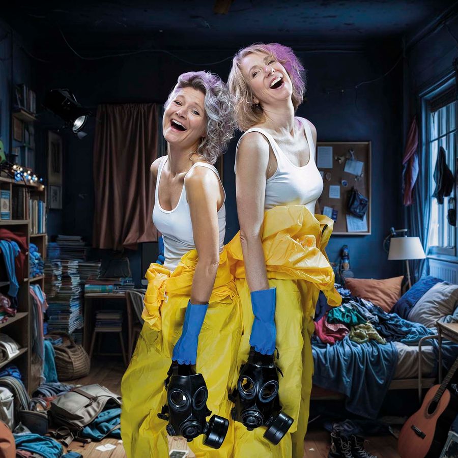 Hanneke en Dianne in gele beschermpakken en blauwe handschoenen, staand in een rommelige kamer. 